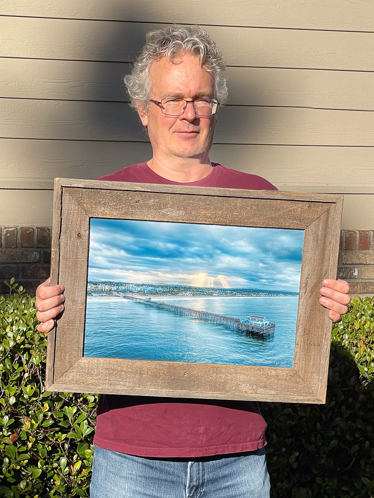 roy holding a photo of the oceanside pier with sun breaking through clouds